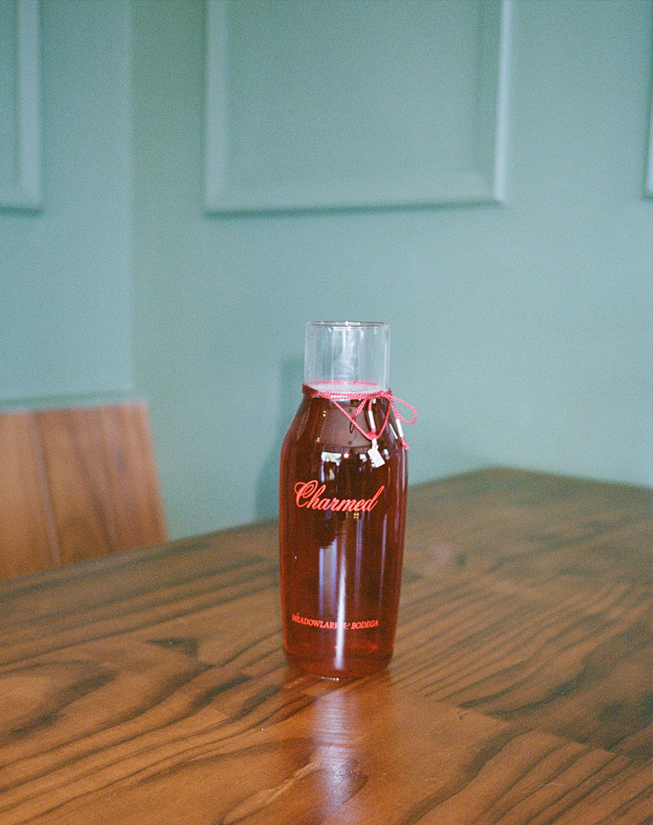 Bottle of 'Charmed' chilled red wine on a wooden table with a light green wall in the background.