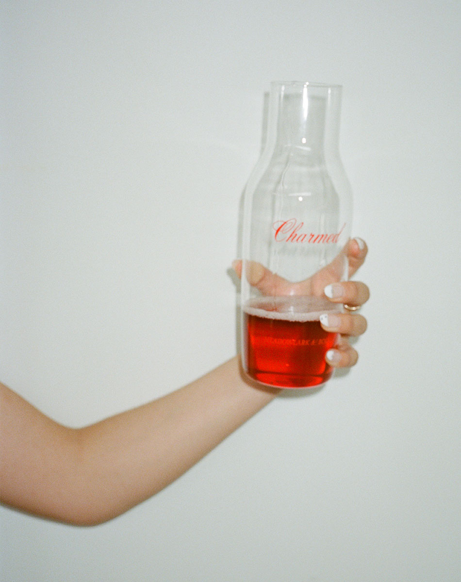 Hand holding a glass carafe with chilled red wine and charmed logo against a plain background