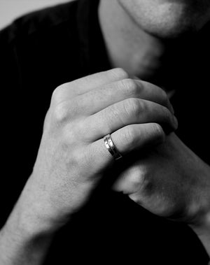 A ring featuring an etched and oxidised dagger design on a band. Worn on a hand.