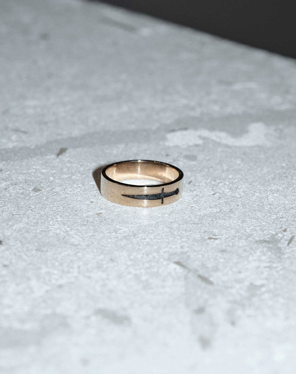 A 9ct yellow gold ring featuring an etched and oxidised dagger design on a band. On stone table.